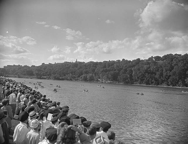 A bird's-eye view of a river with a treeline in the middle ground and people standing as observers in the bottom-left corner.